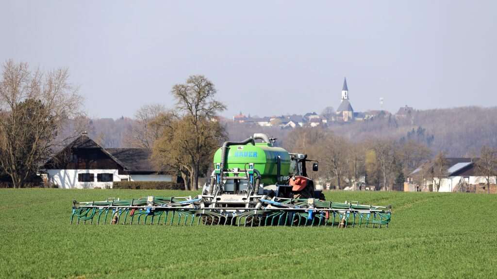 Tracteur en cours d'épandage dans un champ proche d'une ferme. Un village de style européen est visible à l'arrière plan en hauteur.